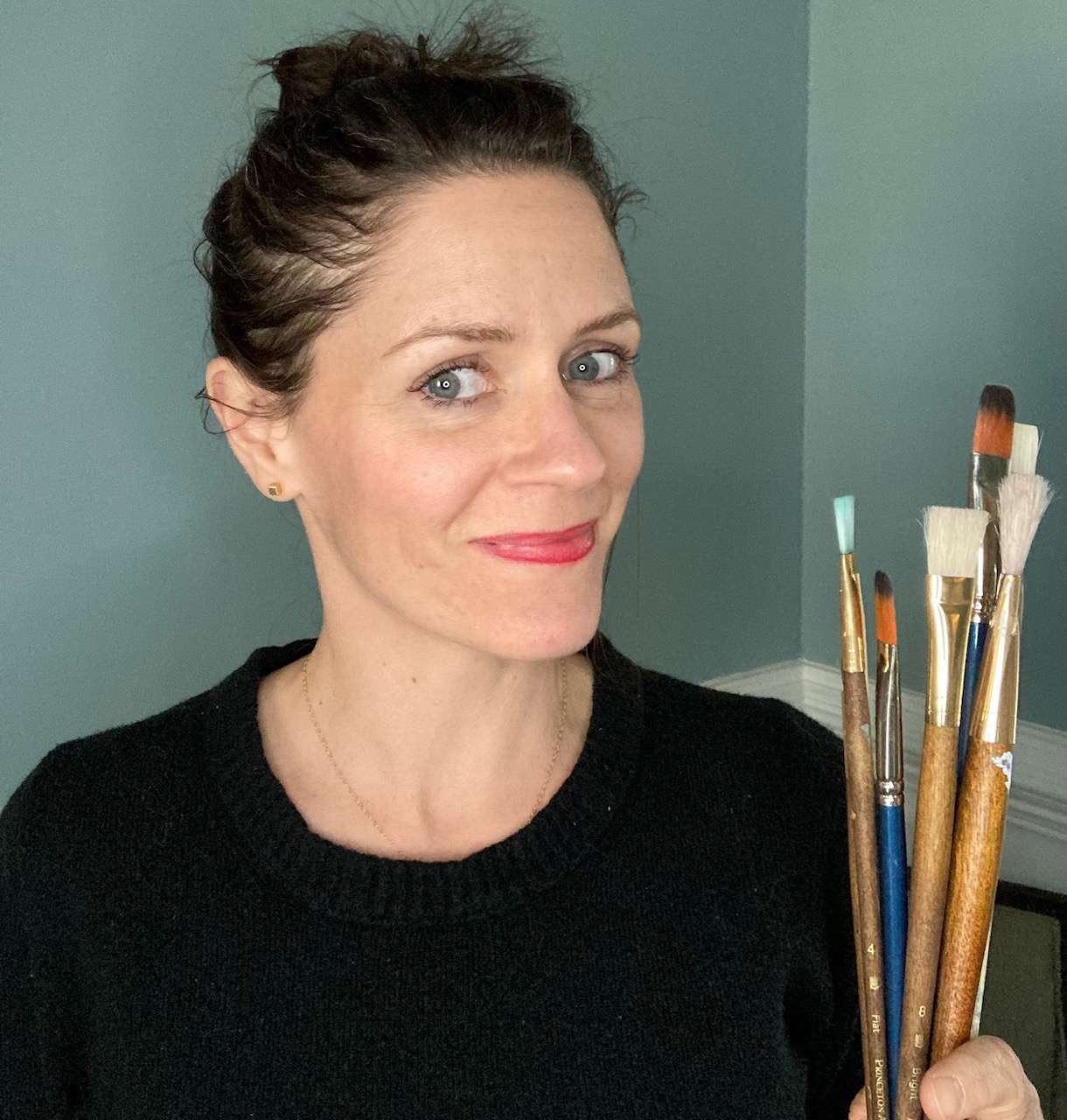 women in black sweater holds several paint brushes while smiling at camera