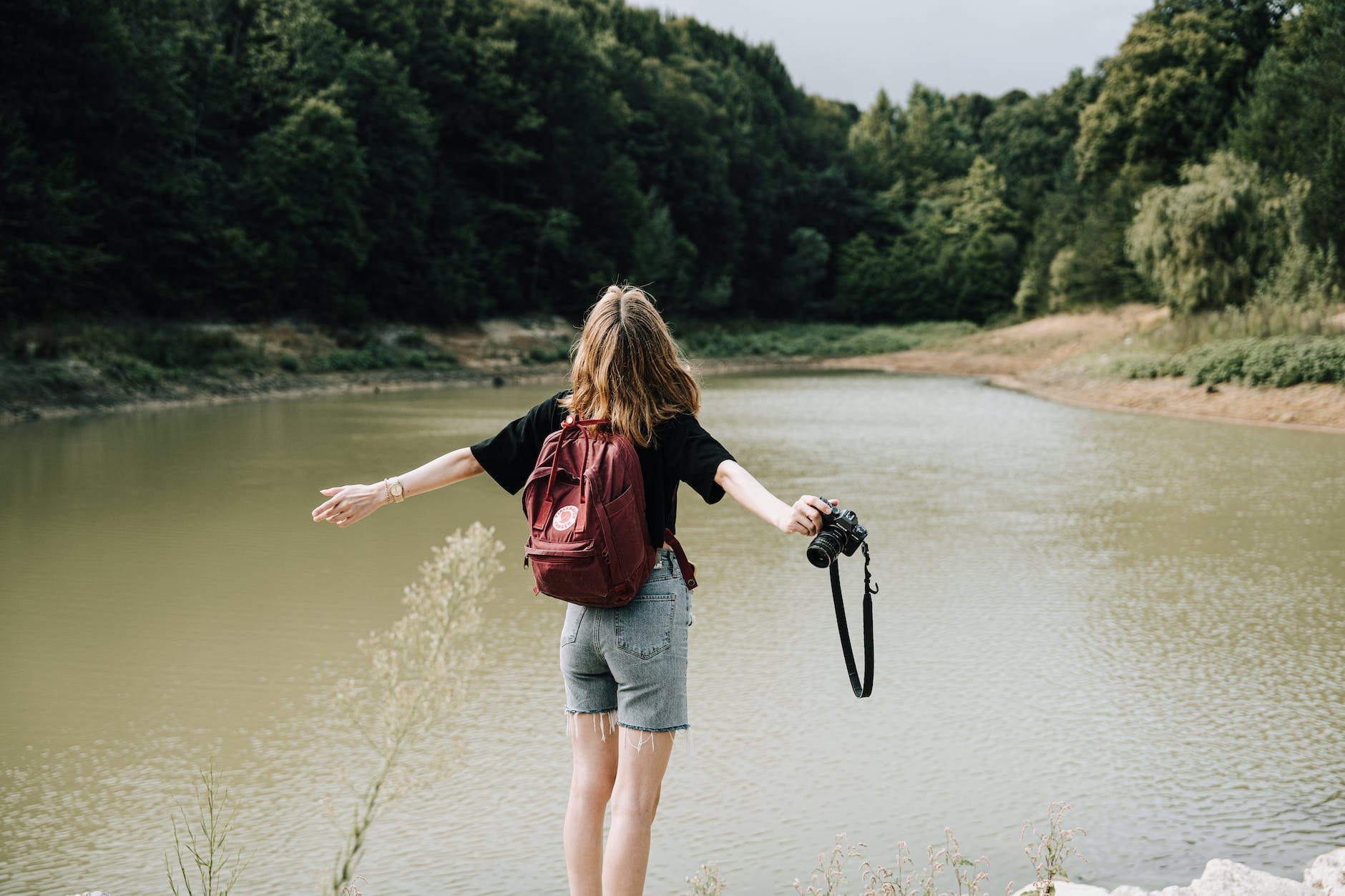 hiker and a camera standing by a lake in the forest with her arms spread